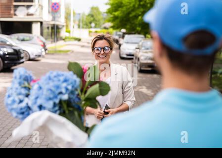 Un jeune liveur en uniforme bleu livre des fleurs à un client de femme Banque D'Images