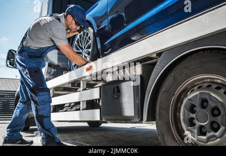 Un travailleur du remorquage qui fournit une voiture neuve du concessionnaire au client. Homme caucasien vérifiant les fixations pour assurer un transport sûr sur son camion de remorquage. Banque D'Images