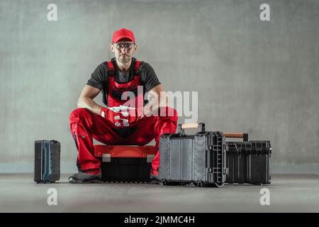 Caucasien Handyman dans l'uniforme de travail rouge assis sur la boîte à outils entouré de plus de boîtes de ses outils de réparation professionnels. Fond gris Uni. Horizo Banque D'Images