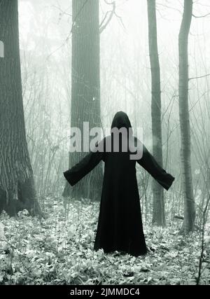 Mort dans un manteau noir à capuchon dans le brouillard de la forêt. Peur de style horreur Banque D'Images