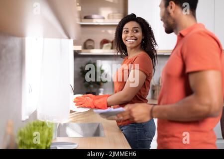 Souriant jeune afro-américain mari et femme dans les mêmes t-shirts laver les assiettes ensemble dans l'intérieur de la cuisine Banque D'Images