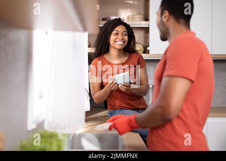 Joyeux jeune famille afro-américaine dans les mêmes t-shirts laver les plats, boire le café et s'amuser dans l'intérieur de la cuisine Banque D'Images