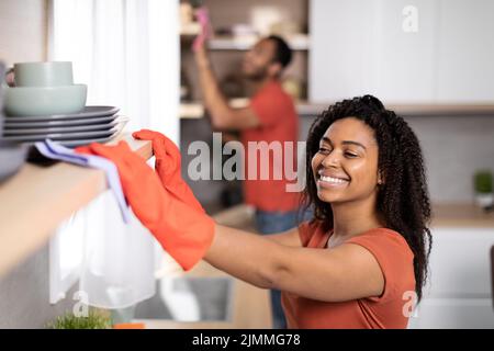 Joyeux jeune couple afro-américain dans le même t-shirts essuie la poussière du mobilier ensemble dans l'intérieur de la cuisine Banque D'Images