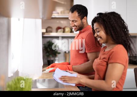 Joyeux homme afro-américain millénaire et dame en même t-shirts laver les plats, profiter de temps ensemble Banque D'Images