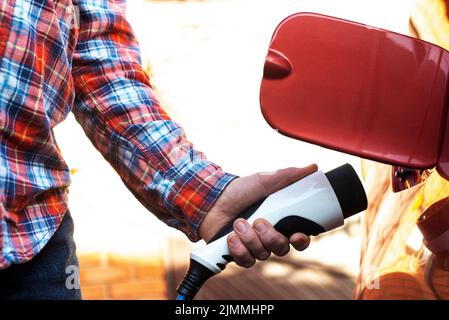 Le modèle mâle se tient près d'une voiture électrique et tient une fiche de charge Banque D'Images