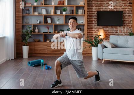 Homme adulte asiatique athlétique faisant une fente, faisant des exercices, travaillant les muscles des jambes à la maison et souriant à la caméra Banque D'Images