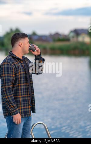 Un jeune homme buvant du café sur une jetée au-dessus d'un lac ou d'une rivière tournant pour admirer la vue Banque D'Images