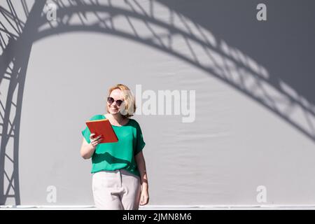 Bonne fille dans des lunettes de soleil parcourant la tablette, s'appuyant sur le mur dans la rue, belle ombre. Formation à distance, formation en ligne et travail Banque D'Images