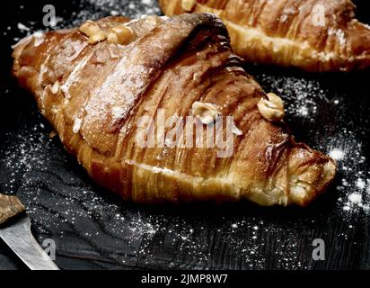 Croissant cuit sur une planche de bois et arrosé de sucre en poudre, table noire. Pâtisseries appétissantes pour le petit déjeuner Banque D'Images