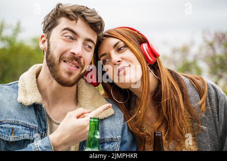 Deux amis heureux partageant un casque pour écouter leur chanson préférée, assis ensemble dans le parc - concept de personnes et de technologie Banque D'Images