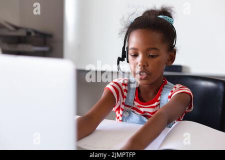 Une écolière afro-américaine élémentaire portant un casque tout en utilisant un ordinateur portable au bureau en classe Banque D'Images