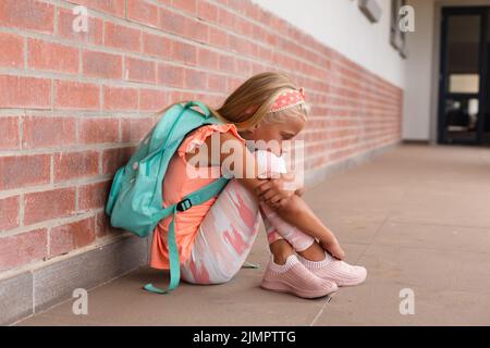 Pleine longueur de triste école élémentaire caucasienne embrassant les genoux tout en étant assis sur le plancher à l'école Banque D'Images