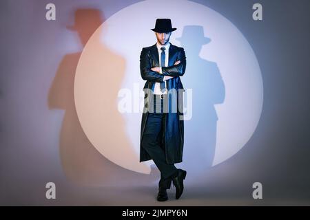 Portrait complet d'un homme doué et confiant en manteau noir et chapeau avec bras croisés posant sur le studio à la lumière du spot. Banque D'Images