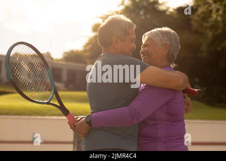 Couple senior biracial romantique et gai se tenant sur un court de tennis au coucher du soleil Banque D'Images