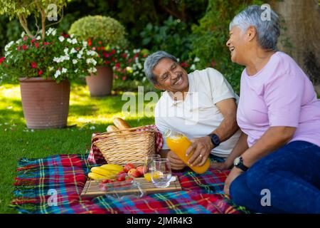 Homme biracial senior souriant versant du jus dans le verre tout en appréciant le pique-nique avec la femme dans le parc Banque D'Images