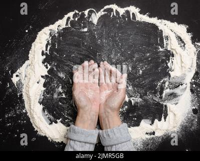 Farine de blé blanc tamisée sur une table noire et deux mains femelles, vue du dessus. Cuisiner à la maison Banque D'Images