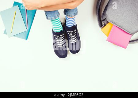 Enfant portant différentes paires de chaussettes. Enfant dans des chaussettes et des baskets colorées mal assorties, assis sur fond blanc, avec sac à dos, livres et école Banque D'Images