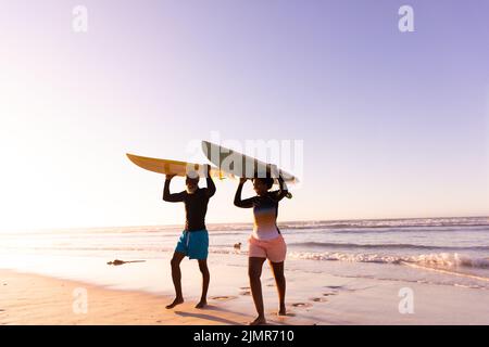 Couple afro-américain transportant des planches de surf sur la tête tout en marchant contre la mer et ciel clair Banque D'Images