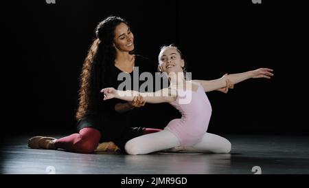 Femme hispanique entraîneur de danse avec les cheveux longs bouclés aider la jeune fille adolescente à se plier le dos tenant les mains sur le côté assis sur le plancher ballerina gymnaste Banque D'Images