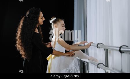 Ado fille enfant adolescent ballerina en tutu écouter conseils de femme danseuse entraîneur enseignant apprendre à danser dans l'école de danse ballet barre s'étirant de la hanche Banque D'Images