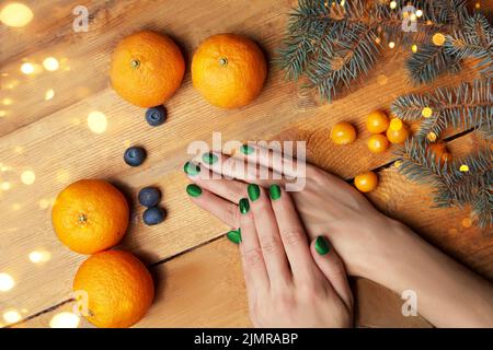 Mains de femmes avec manucure verte couchée sur la table, vue de dessus. Confort à la maison, mandarines, branche de sapin sur le fond des lumières de Noël. Photo de haute qualité Banque D'Images