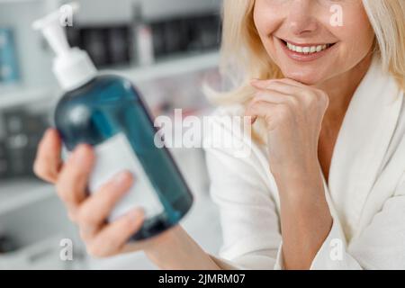 Belle femme blonde senior souriante est assise dans le centre de bien-être et tient une bouteille Banque D'Images