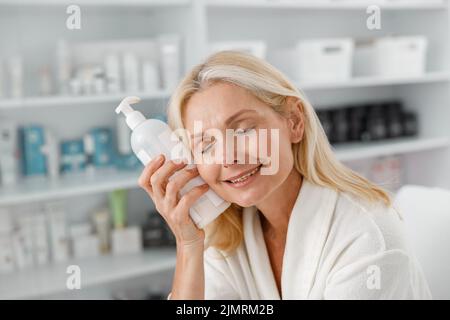 Femme âgée souriante assise dans un centre de bien-être contenant une bouteille de produit de beauté Banque D'Images