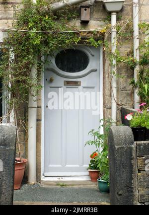 Ancienne porte de maison traditionnelle en bois peint en gris bleu avec boîte aux lettres en laiton et fenêtre avec un cadre blanc entouré de fleurs, pot Banque D'Images