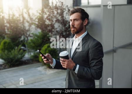 Homme d'affaires attrayant avec téléphone et café debout près du bâtiment de bureau et vue loin Banque D'Images