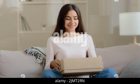 Surprise arabe latino femme satisfait client shopaholic s'assoit sur le canapé à la maison livraison ouverte colis boîte postale carton déballer présent cadeau regarder Banque D'Images