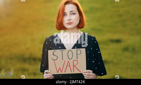 Malheureux caucasien jeune femme redhead activiste tenir démontrant bannière avec l'inscription arrêter la guerre triste jolie fille stand dehors dans le champ show Banque D'Images