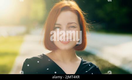 Portrait de la femme heureux beau visage rouge fille élégante avec la coiffure courte souriant posant dans la ville rayons solaires envoie l'air baiser par geste de main Banque D'Images