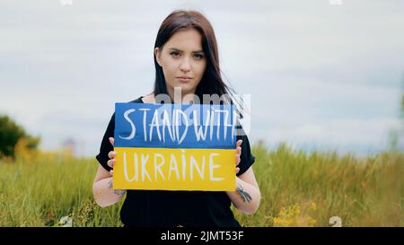 Une femme dépressive bouleversée élève une bannière d'art carton avec l'inscription politique stand avec l'Ukraine manifestant protestant coloré jaune bleu Banque D'Images