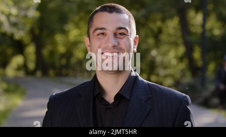 Homme sourire portrait à l'extérieur adulte homme homme stand à l'extérieur porte costume noir rencontrer quelqu'un dans le parc regarder l'appareil photo vous dit bonjour main vague gai Banque D'Images