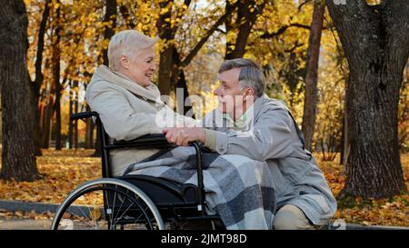 Homme âgé caucasien tenant une femme handicapée à la main, le mari en fauteuil roulant accompagne la femme malade pendant la réhabilitation dans le parc d'automne Banque D'Images