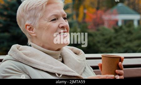 Heureuse grise-cheveux rêveuse femme âgée mature grand-mère boire du café thé chaud de la tasse jetable assis sur le banc de parc senior sain gai Banque D'Images