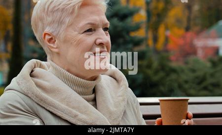 Heureuse rêveuse grise-cheveux âgés mature femme grand-mère assis sur le banc du parc boire du café thé chaud de la tasse jetable sain gai senior Banque D'Images