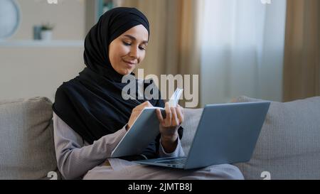 Gaie jeune musulmane arabe dame jeune femme assis sur un canapé confortable femme journaliste indépendant prendre des notes dans le journal notant l'information écrire livre tâches quotidiennes Banque D'Images