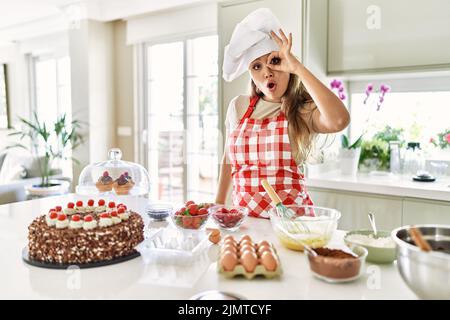 Belle jeune brunette pâtisserie chef femme cuisant des pâtisseries à la cuisine faisant ok geste choqué avec surprise visage, oeil regardant à travers les doigts. Banque D'Images