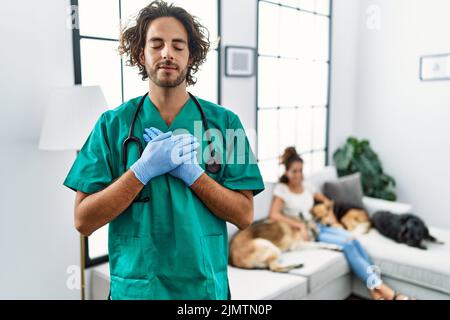 Jeune vétérinaire homme vérifiant les chiens à la maison souriant avec les mains sur la poitrine avec les yeux fermés et geste reconnaissant sur le visage. Concept de santé. Banque D'Images