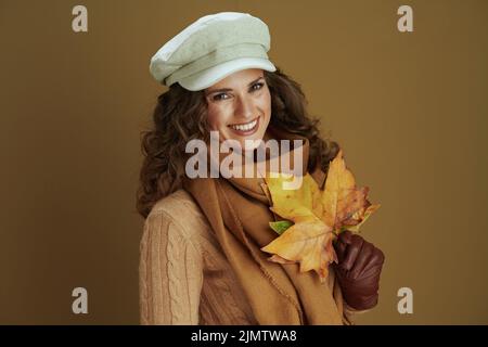 Bonjour septembre. Portrait d'une femme moderne d'âge moyen souriante en écharpe avec gants en cuir et feuilles d'érable jaune d'automne isolées sur fond de bronze Banque D'Images