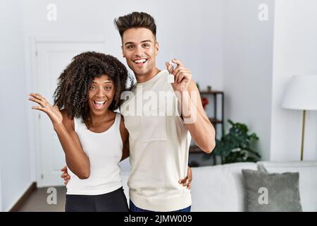 Jeune couple interracial tenant les clés de la nouvelle maison célébrant la victoire avec le sourire heureux et l'expression du gagnant avec les mains levées Banque D'Images