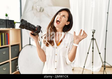 Belle femme caucasienne travaillant en tant que photographe au studio de photographie hurlant et fou et hurlant avec l'expression agressive et les bras levés. Banque D'Images