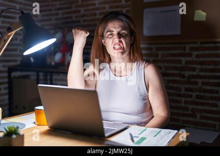 Brunette femme travaillant au bureau la nuit en colère et fou levant poing frustré et furieux tout en criant avec colère. Rage et agressif concept Banque D'Images