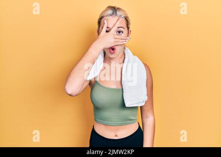 Jeune fille blonde portant des vêtements de sport et une serviette en choc couvrant le visage et les yeux avec la main, regardant à travers les doigts avec l'expression embarrassée. Banque D'Images