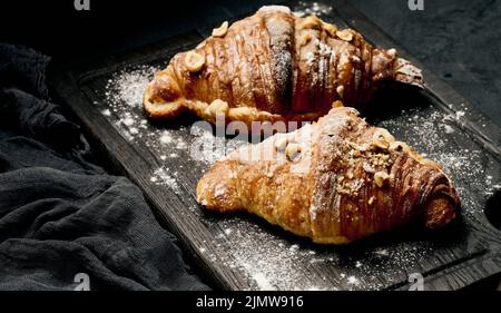 Croissant cuit sur une planche de bois et arrosé de sucre en poudre, table noire. Pâtisseries appétissantes pour le petit déjeuner Banque D'Images