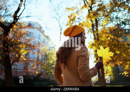 Bonjour automne. Vue de derrière une femelle en manteau beige et chapeau orange avec des feuilles jaunes d'automne dehors dans la ville en automne. Banque D'Images