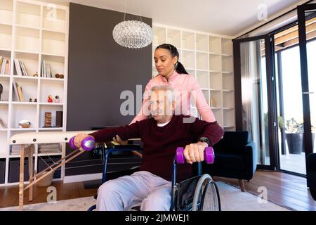 Une physiothérapeute biraciale aide un homme âgé caucasien à lever des haltères à la maison Banque D'Images