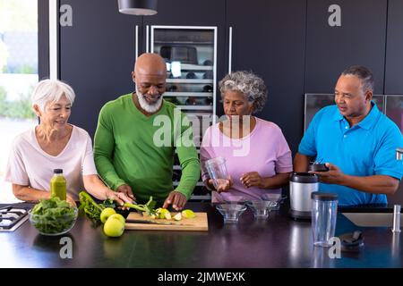 Amis séniors multiraciaux qui font du smoothie avec des pommes Granny smith et des légumes-feuilles dans la cuisine Banque D'Images