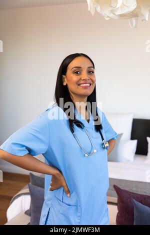 Portrait d'une femme kinésithérapeute biracial souriante avec des bras akimbo debout contre le mur à la maison Banque D'Images